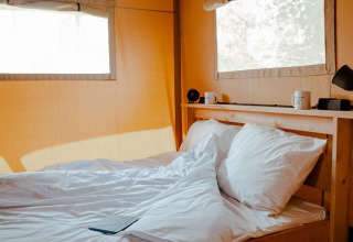 Cama acogedora con sábanas blancas en una tienda de glamping iluminada y ventanas grandes de lona clara.