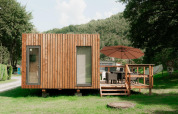 A modern wood-clad glamping cabin with a deck, patio umbrella, and chairs, set in a green, wooded area.