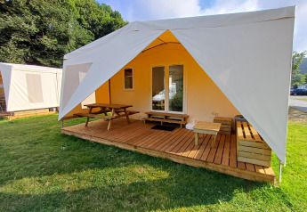 Safari tent with wooden deck, benches, and picnic table, set outdoors on green grass in a camping area.