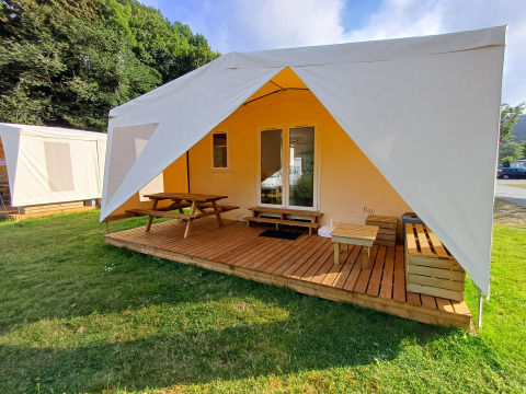 Tienda de safari con terraza de madera, bancos y mesa de picnic al aire libre en una zona de césped verde.