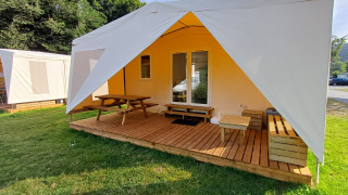 Safari-Zelt mit Holzterrasse, Sitzbank und Picknicktisch im Freien auf einer Wiese an sonnigem Morgen.