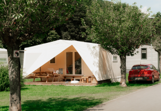 Safaritent met veranda, picknicktafel en stoelen naast een rode auto, omgeven door bomen en gras.