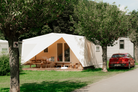 Tienda safari con porche cubierto, mesa de picnic y coche rojo cerca, rodeada de árboles y césped.