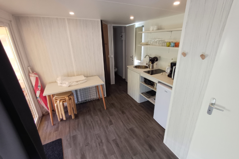 Interior de tienda safari con zona de cocina, frigorífico pequeño, mesa, taburetes y estantes con vajilla.