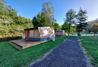 Foto di una tenda safari con veranda in legno su un campeggio, circondata da erba e alberi verdi.