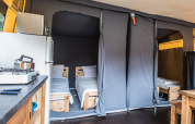 Interno di una tenda safari con due letti singoli, angolo cottura, frigorifero e tavolo in legno.