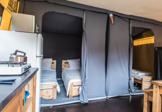 Interno di una tenda safari con due letti singoli, angolo cottura, frigorifero e tavolo in legno.