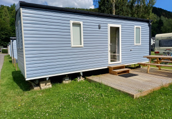 Modern blue glamping cabin with small steps, wooden deck, and picnic table on green grass outdoors.