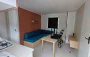 Sistemazione glamping moderna con divano blu, tavolo da pranzo, sedie e piccolo angolo cottura.