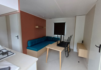 Sistemazione glamping moderna con divano blu, tavolo da pranzo, sedie e piccolo angolo cottura.