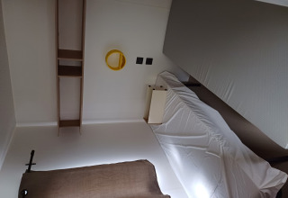 Una camera di glamping con letto singolo, lenzuolo bianco, mensole, specchio tondo giallo e tenda marrone.