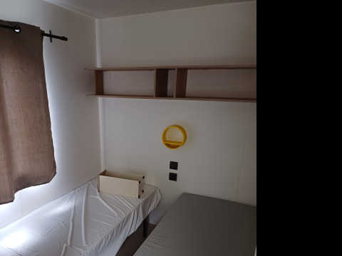 Una camera di glamping con letto singolo, lenzuolo bianco, mensole, specchio tondo giallo e tenda marrone.