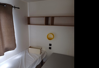 Una camera di glamping con letto singolo, lenzuolo bianco, mensole, specchio tondo giallo e tenda marrone.