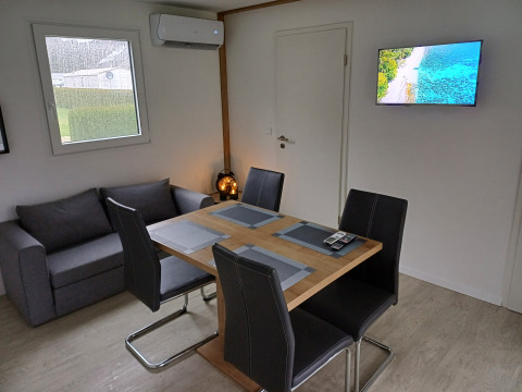 Moderne glampingaccommodatie met eettafel, vier stoelen, grijze zetel, airco en tv aan de muur.