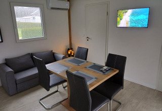Alloggio glamping moderno con tavolo da pranzo, quattro sedie, divano grigio, aria condizionata e TV a muro.