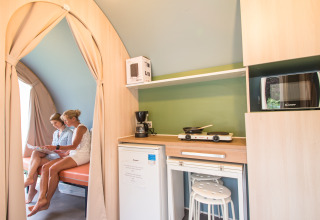 Vista interior de la tienda Coco Sweet en Camping La Colline, Bélgica, con dos mujeres, cocina y zona de estar.
