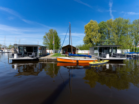 Dos modernos barcos casa y un velero naranja en Marina Heeg, Países Bajos, rodeados de agua y árboles.