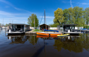 To moderne husbåde og en orange sejlbåd ved Marina Heeg i Holland, omgivet af vand og grønne træer.