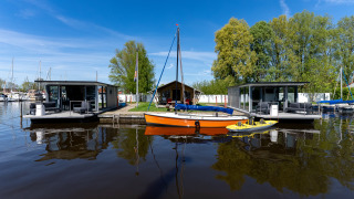 Twee moderne woonboten en een oranje zeilboot bij Marina Heeg in Nederland, omgeven door water en bomen.