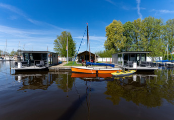 To moderne husbåde og en orange sejlbåd ved Marina Heeg i Holland, omgivet af vand og grønne træer.