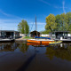 Twee moderne woonboten en een oranje zeilboot bij Marina Heeg, Nederland, met water en groene bomen.