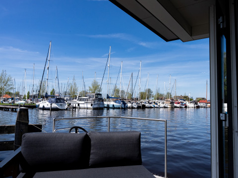 Blick von Houseboat Heeg in Marina Heeg, Niederlande, mit Sofa und Segelbooten bei blauem Himmel.