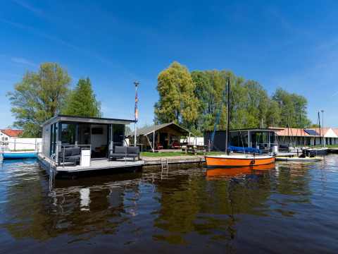 Casa flotante en Marina Heeg, Países Bajos, atracada junto a barcos y rodeada de árboles y naturaleza.