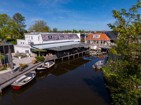 Casa flotante en Marina Heeg, Países Bajos, con terraza junto al canal y botes bajo un cielo azul claro.