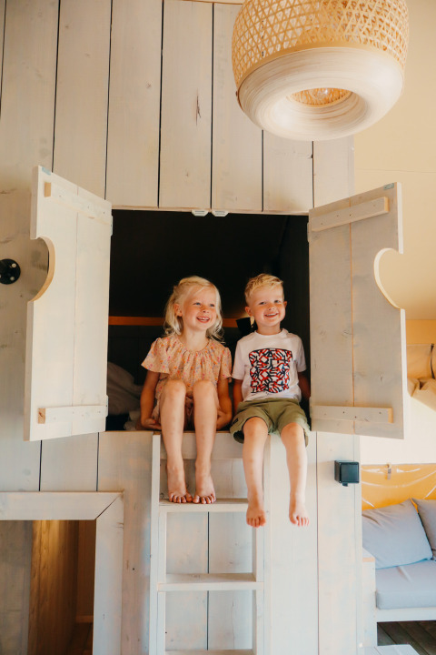 Zwei fröhliche Kinder sitzen in einem Bett mit offenen Türen in einem Safari-Zelt namens Safari cottage.