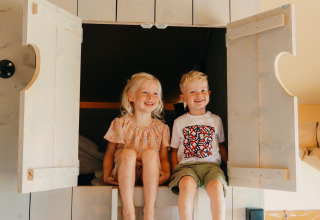 To glade børn sidder i en seng med åbne døre i et safari telt, Safari cottage, med lyst træinteriør.