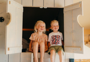 To glade børn sidder i en seng med åbne døre i et safari telt, Safari cottage, med lyst træinteriør.