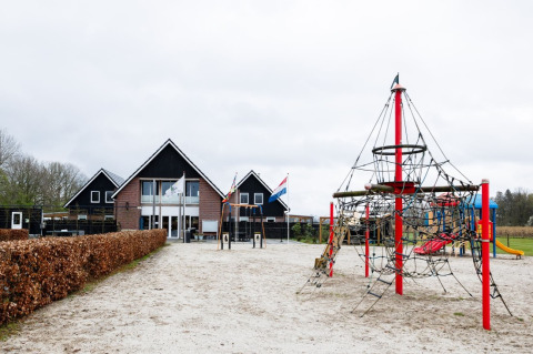 Spielplatz mit Klettergerüst und Rutsche vor einem Ferienparkgebäude in Drenthe, Niederlande, bei bewölktem Himmel.