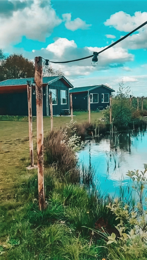 Idyllisk landskab ved The Kremmer feriepark i Drenthe med hytter, dam og dekorative lyskæder.