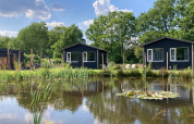 To sorte hytter ved en dam omgivet af grønt i The Kremmer feriepark, Drenthe, Holland på en solskinsdag.