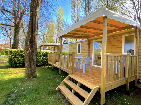 Holzchalet mit überdachter Terrasse und Gartenmöbeln auf Camping Le Céou, umgeben von Bäumen und Grün.