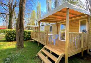 Wooden chalet with covered veranda and outdoor seating at Camping Le Céou, surrounded by lush greenery.
