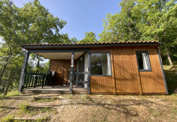 Cabaña glamping de madera con porche cubierto y muebles de exterior, rodeada de árboles verdes.