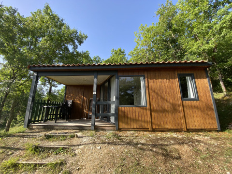 Houten glampinghuisje met overdekte veranda en tuinmeubilair, omringd door groene bomen.