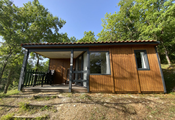 Houten glampinghuisje met overdekte veranda en tuinmeubilair, omringd door groene bomen.
