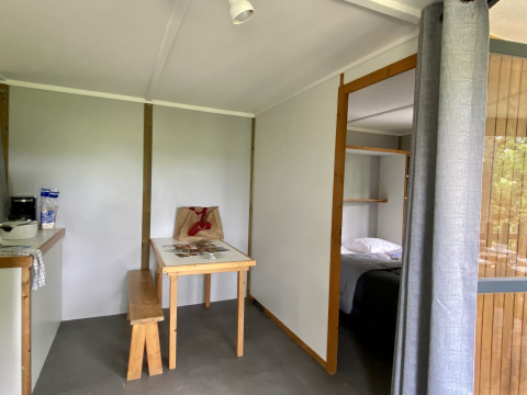 Interieur van een glampingverblijf met tafel, bank, kitchenette en zicht op de slaapkamer.