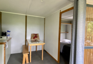 Interieur van een glamping-accommodatie met tafel, bank, keukenhoek en zicht op de slaapkamer.