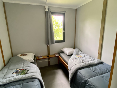 Glamping room with two single beds, grey bedding, a central window, and magazines placed on the duvets.