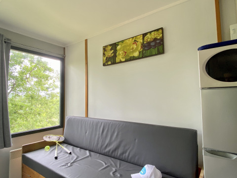 Comfortabele glampingruimte met grijze zetel, groot raam, microgolf, koelkast en kunstwerk aan de muur.