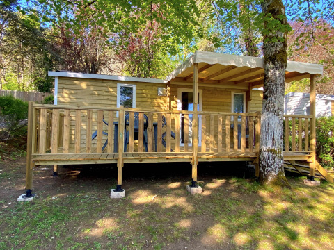 Stacaravan houten chalet met overdekt terras, gelegen tussen bomen en voorzien van zitplaatsen buiten.