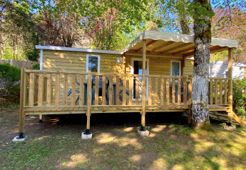 Stacaravan Holzhütte mit überdachter Veranda in grüner Umgebung und Sitzgelegenheiten draußen.