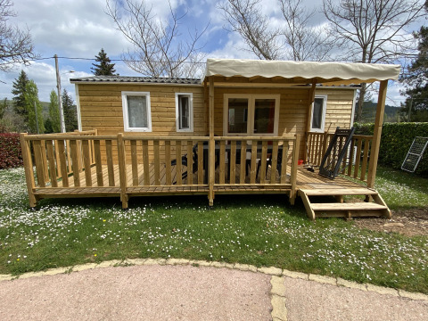 Wooden cabin with covered porch and steps, surrounded by green lawn and flowers under a cloudy sky.