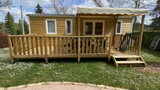 Houten chalet met overdekt terras en trapje, omringd door gras en bloemetjes onder een bewolkte hemel.