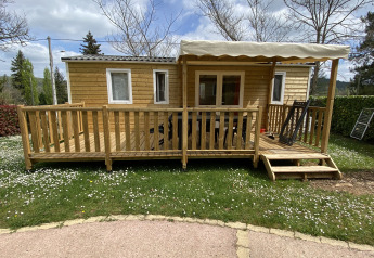Houten chalet met overdekt terras en trapje, omringd door gras en bloemetjes onder een bewolkte hemel.