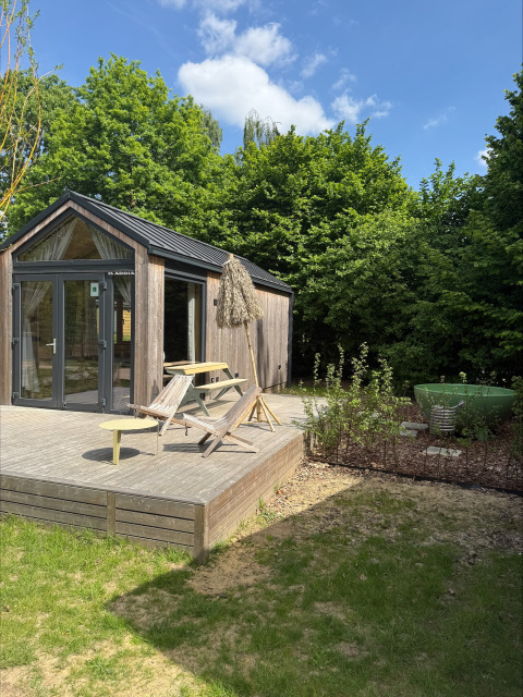 Pequeña casa de madera con terraza, mobiliario exterior y dutchtub, rodeada de árboles en Urban-Gardens, Bélgica.