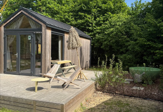 Pequeña casa de madera con terraza, mobiliario exterior y dutchtub, rodeada de árboles en Urban-Gardens, Bélgica.